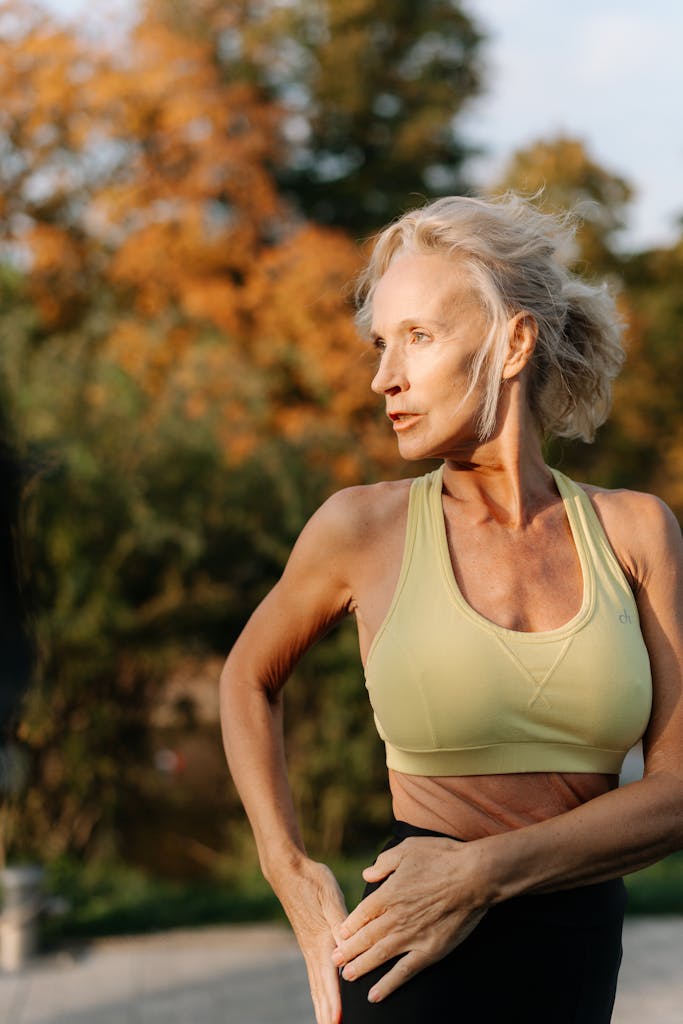 Elderly woman in sports bra practicing yoga outdoors with autumn foliage in background.