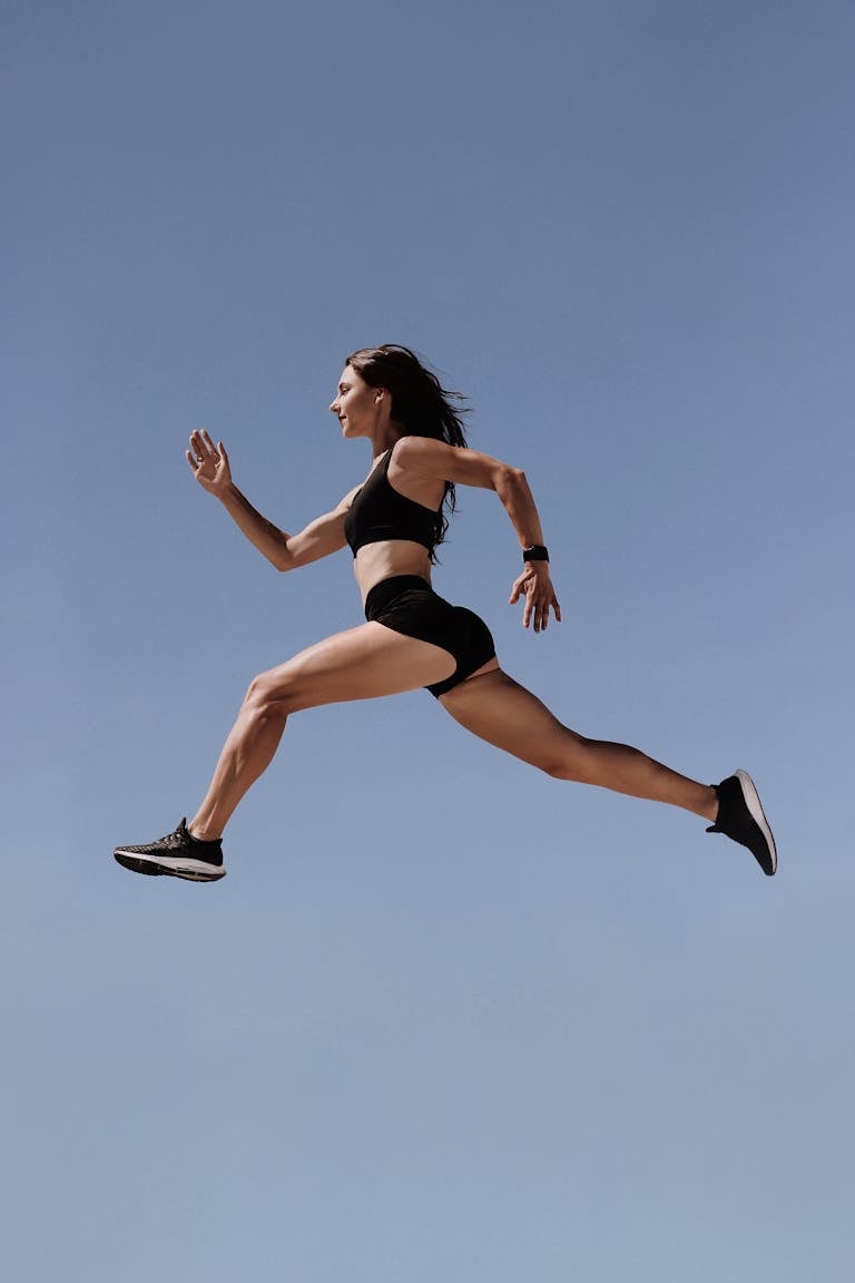 Energetic woman in athletic wear leaping against clear blue sky, symbolizing freedom and fitness.