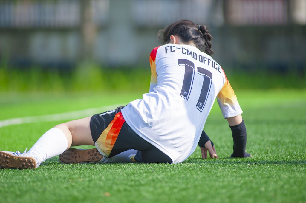 Female soccer player in sports uniform resting on green field in sunlight.