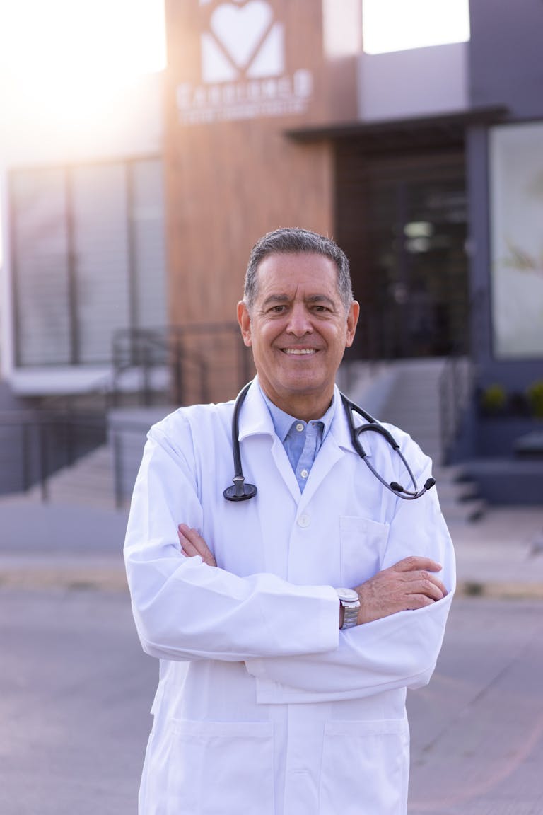 Smiling doctor in lab coat with stethoscope, standing outdoors.