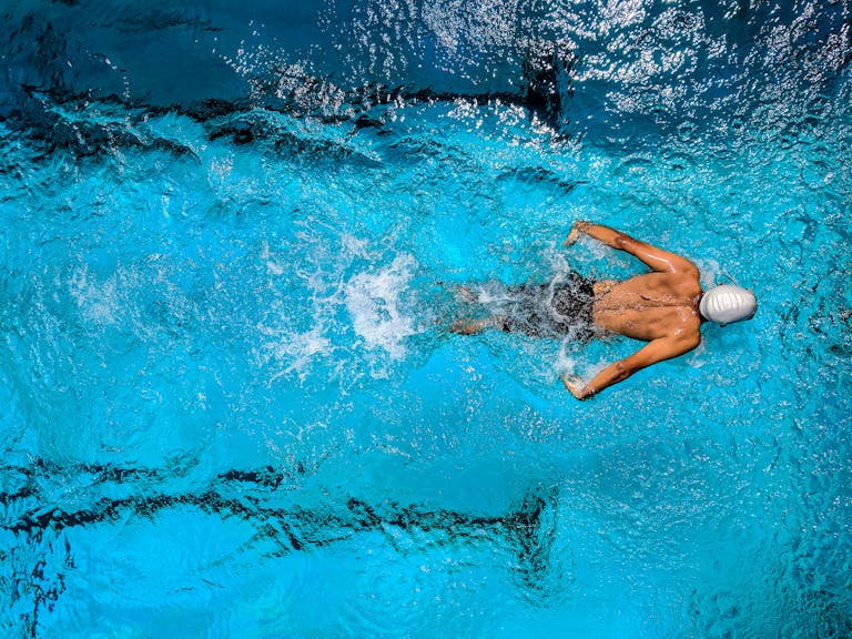 Top view of a swimmer wearing a cap, performing a front crawl stroke in a clear blue swimming pool.